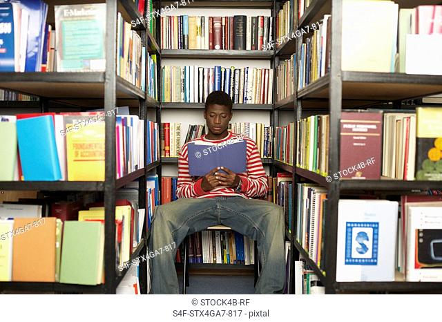 Student sitting between bookshelves while reading a book