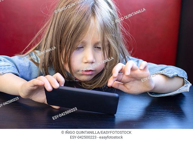Portrait of five years old child with blue denim jacket, sitting in red sofa and black table in a restaurant, touching with finger a smartphone mobile in her...