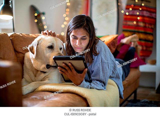 Labrador retriever and young woman lying on living room sofa looking at digital tablet