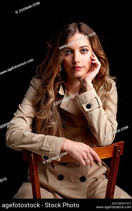 Portrait of young beautiful blonde woman is sitting on a chair. Female in beige cloak on black background