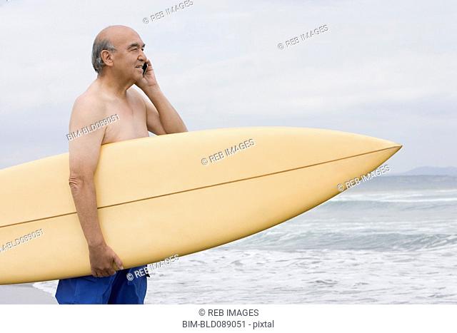 Senior Hispanic man holding surfboard on beach and talking on cell phone