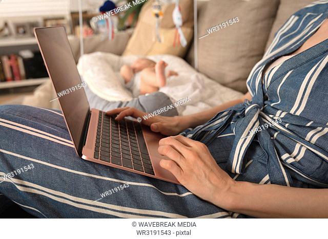 Mother working on laptop baby lying in crib besides her at home
