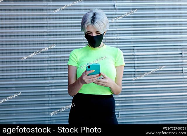 Blond woman standing in front of metal background, wearing face mask, using smartphone