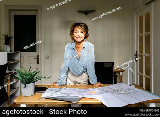 Smiling female architect leaning on desk while standing in office