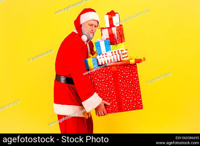 Portrait of tired elderly man with gray beard wearing santa claus costume carrying many boxes with presents, preparing for Christmas celebrations