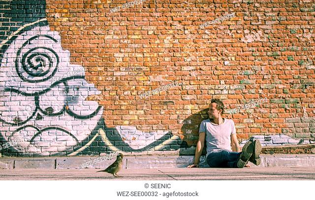 USA, New York City, man sitting at brick wall with graffiti and sparrow in foreground