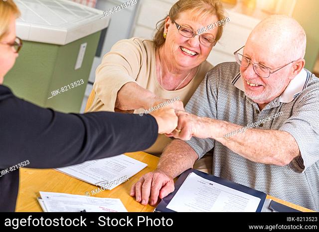 Senior adult couple celebrating with fist bump over documents in their home with agent at signing