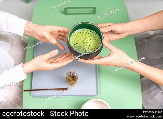 Pair of girls holding an emerald bowl with matcha green tea in their hands over the green metallic table indoors. On the table there is light bowl