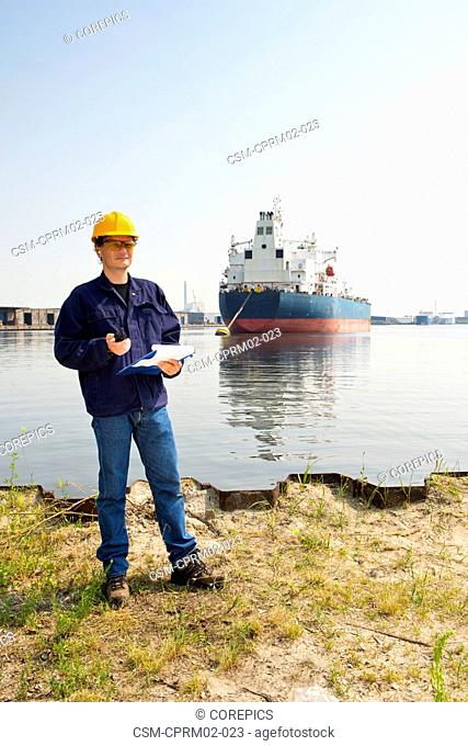 Docker posing on the water's edge with a clipboard and cb radio in his hand, in front of a large oil tanker, moored off in Amsterdam Harbor