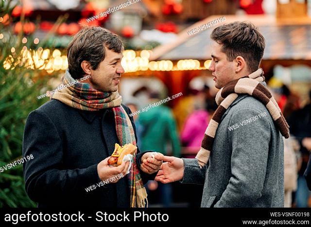 Smiling father holding hot dog and talking with son standing at Christmas market