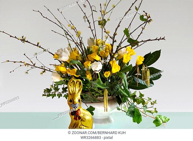 Osterstrauss mit gelben Osterglocken, weissen Tulpen, Zweigen und Efeuranken in einer weissen Terrine auf einem Glastisch