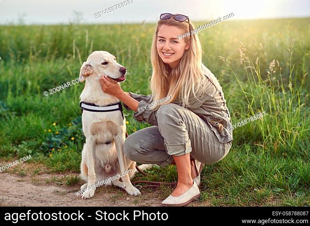 Happy smiling golden dog wearing a walking harness sitting facing its pretty young woman owner who is caressing it with a loving smile outdoors in countryside