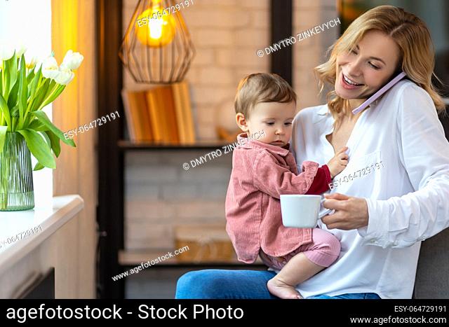 Mother talking on telephone holding baby and drinking coffee, enjoying free time with daughter and hot beverage