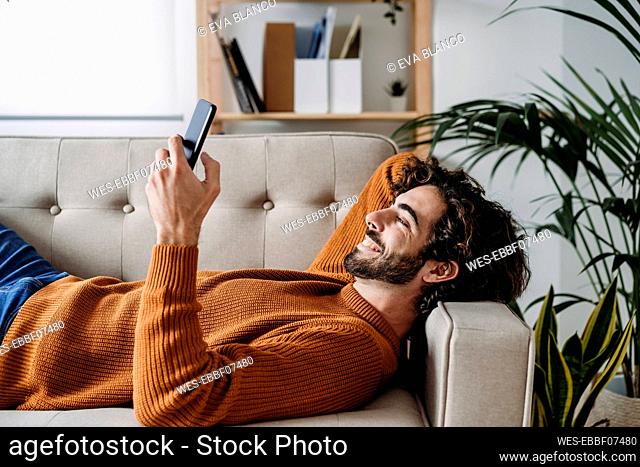 Happy young man using smart phone relaxing on sofa at home