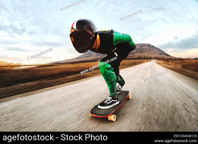 A young man in a fullface helmet and a leather suit in a special rack rides a longboard on afsaltu sunset in the background mountains and beautiful sky at high...