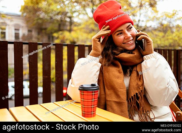 Smiling woman looking away while talking on smart phone sitting at sidewalk cafe