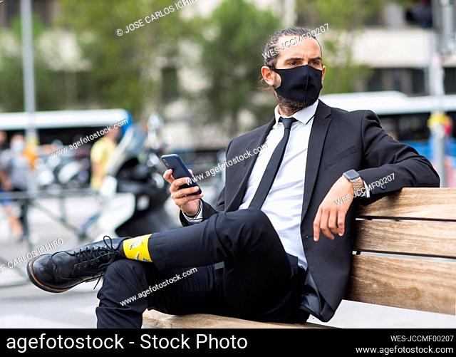 Portrait of businessman wearing protective face mask sitting on bench with smart phone in hand