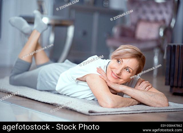 A Woman Over 50 Enjoys Relaxation After Yoga. She Lies on the Floor at Home. Woman Gently Smiles. Yoga Gives Her Pleasure. Close Up Shot