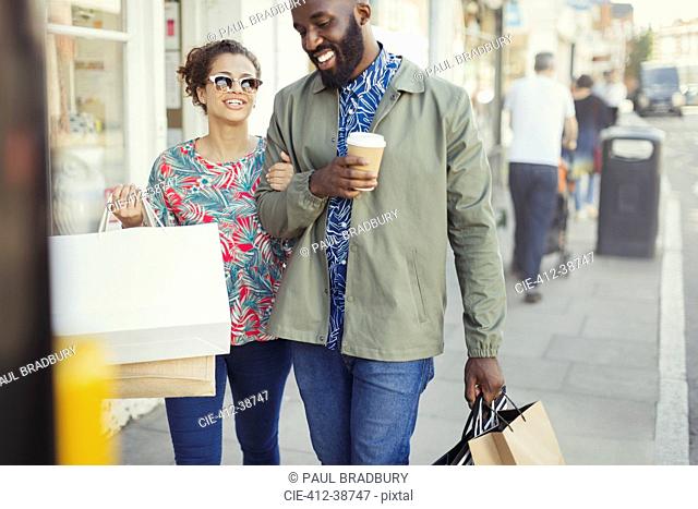 Smiling young couple with coffee and shopping bags walking along storefront