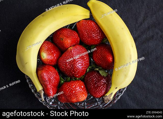 Large strawberries in a glass bowl complemented by two bananas. Still life, macro, black background
