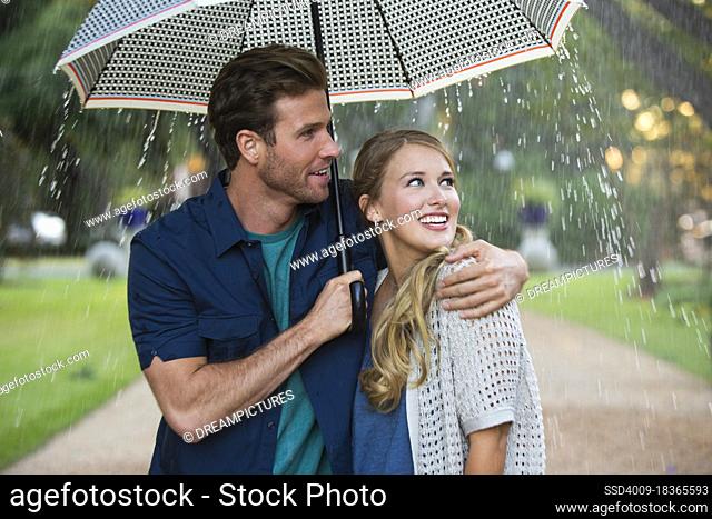 Young couple walking through park with umbrella in the rain, both looking out at the falling rain