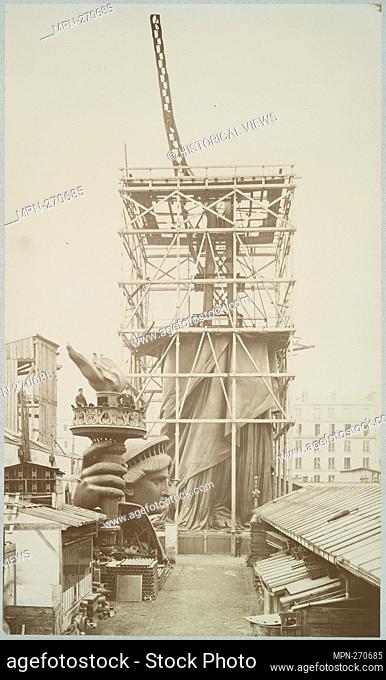 Assemblage of the Statue of Liberty in Paris, showing the bottom half of the statue erect under scaffolding, the head and torch at its feet