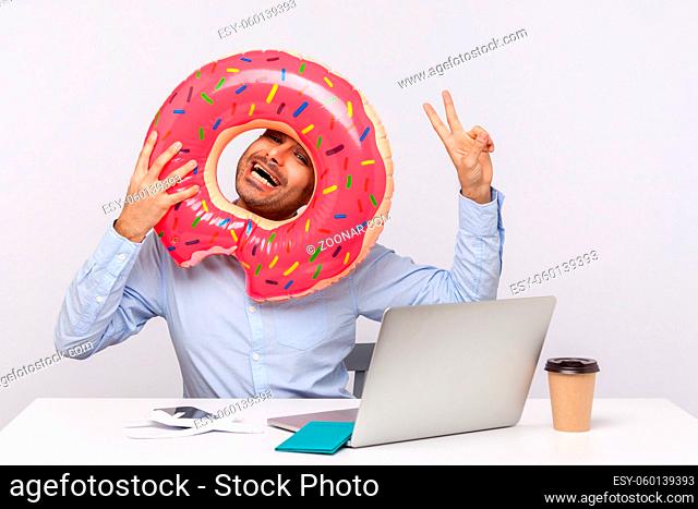 Extremely joyful man looking through rubber ring and showing victory gesture, sitting in office workplace with passport on desk