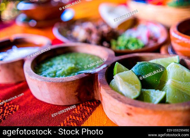 Close-up of clay bowls of lime slices and hot green and red sauces with blurry chopped meat as background. Condiments and sliced vegetables above orange and red...