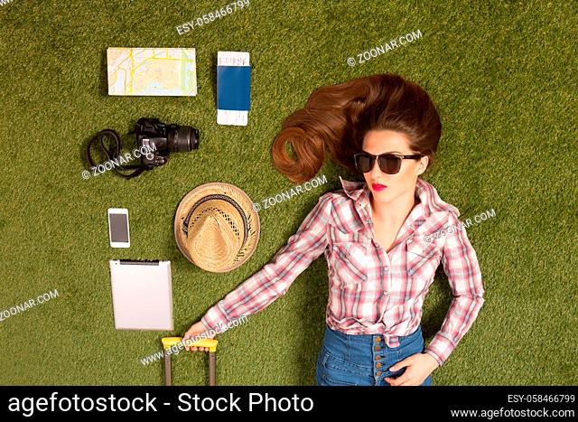 Portrait of beautiful tourist lady in sunglasses lying on green grass among mobile or smart phone, passport, cap, map or guide, etc