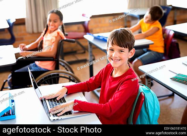 Portrait of caucasian boy with laptop smiling while sitting on his desk at elementary school