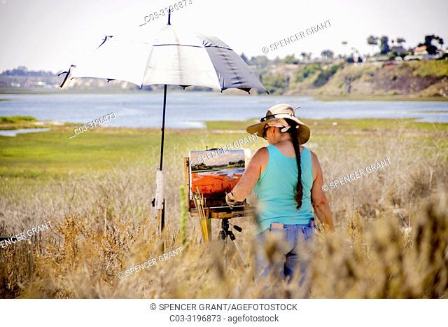Under an umbrella in the sunshine, a female artist works on her plein air outdoor painting that matches her scenic view of Newport Beach, CA