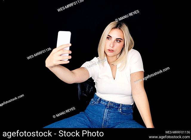 Blond woman taking selfie through smart phone over black background
