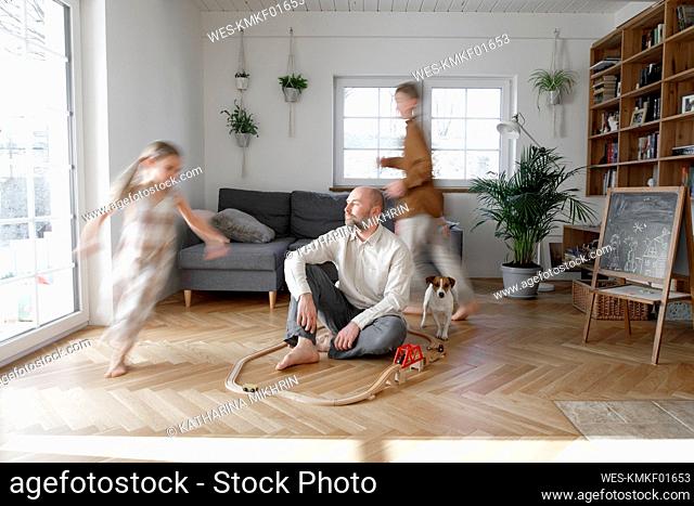 Siblings running around father sitting on floor in living room at home