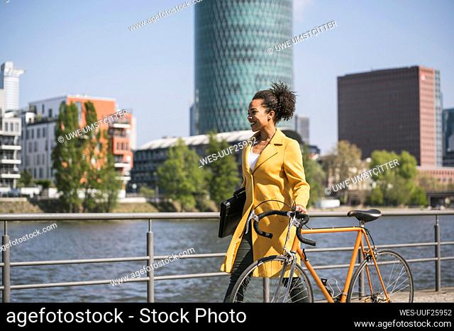 Smiling businesswoman wheeling bicycle by River Main on sunny day