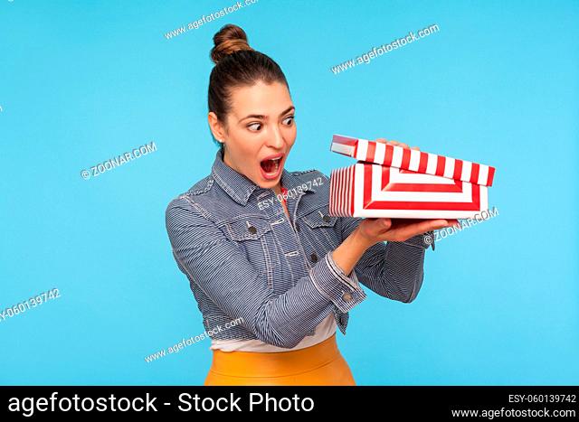 Astonished curious woman with hair bun in denim jacket unpacking opening small carton box, looking inside with surprised amazed expression, shocked by gift