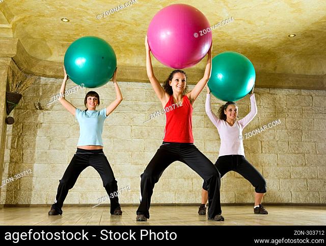 A group of women making exercise holding big balls over heads and standing astride. They're smiling. Low angle view