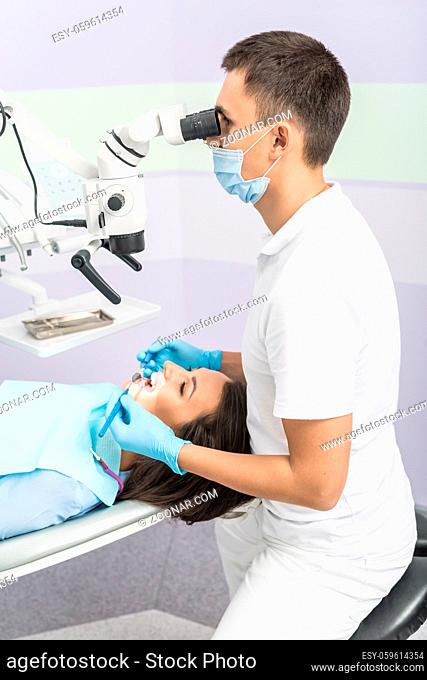 Nice girl with patient bib on a dental chair and dentist who sits next to her. He looks on her teeth using dental microscope and holds dental bur and mirror