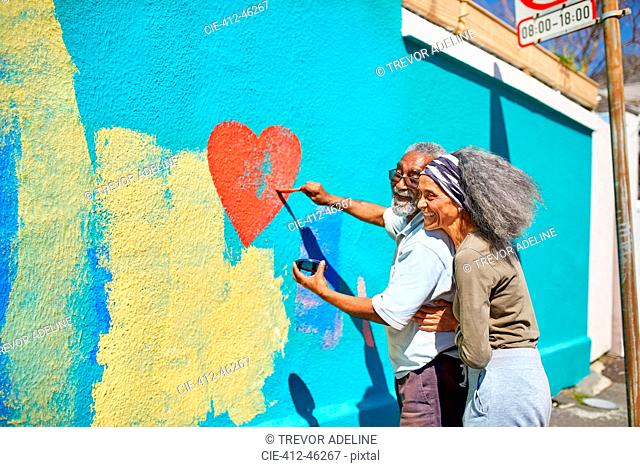 Happy senior couple painting heart-shape mural on sunny wall