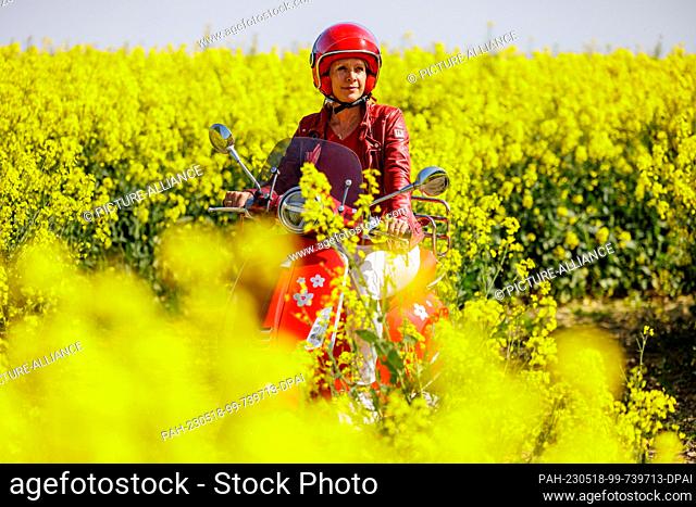 18 May 2023, Schleswig-Holstein, Altenholz: A woman rides her motorized scooter in bright sunshine along a country lane that leads through a field of rape in...