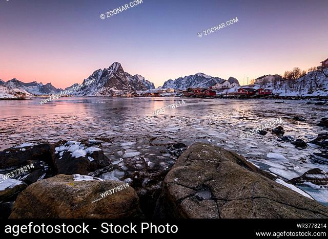 The small fishing village Reine on the Lofoten islands in Norway in winter with steep snowcapped mountains and frozen lake during sunrise