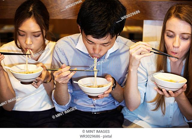 High angle view of three people sitting sidy by side at a table in a restaurant, eating from bowls using chopsticks