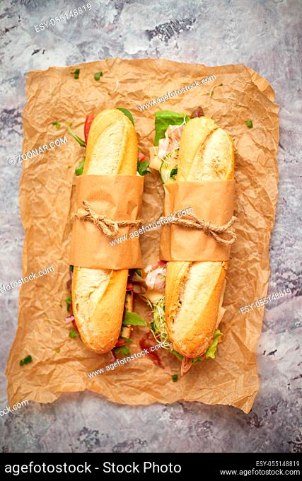 Sandwiches with ham, fresh vegetables and herbs served on brown baking paper over white concrete backdrop, top view