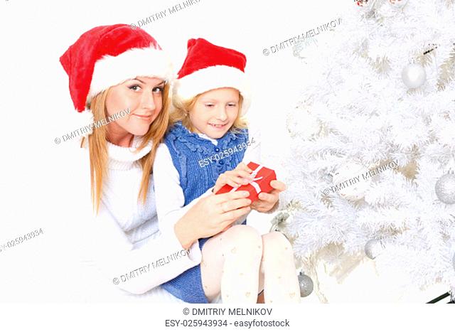 The pretty girl and her mother with gift box are sitting near Christmas tree on a white background