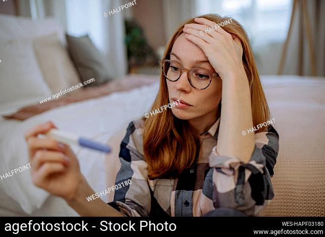 Worried woman wearing eyeglasses sitting with pregnancy testing kit in front of bed at home
