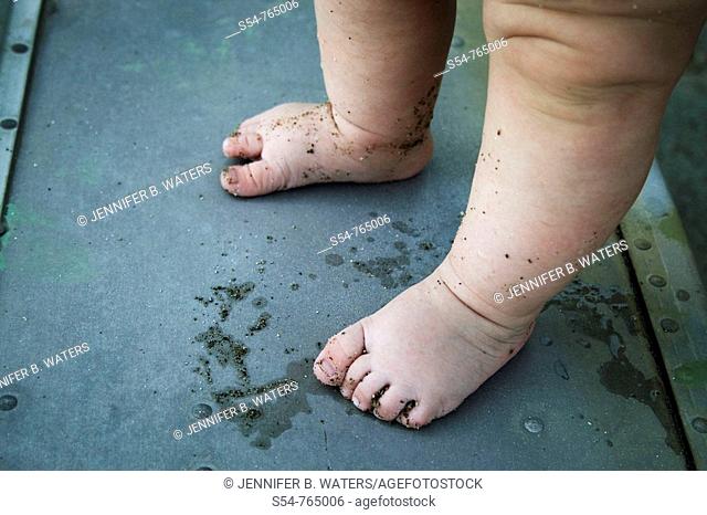 A caucasian baby stands in a boat, with sand on his feet