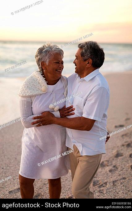 Smiling senior biracial couple looking at each other while standing on shore at beach during sunset
