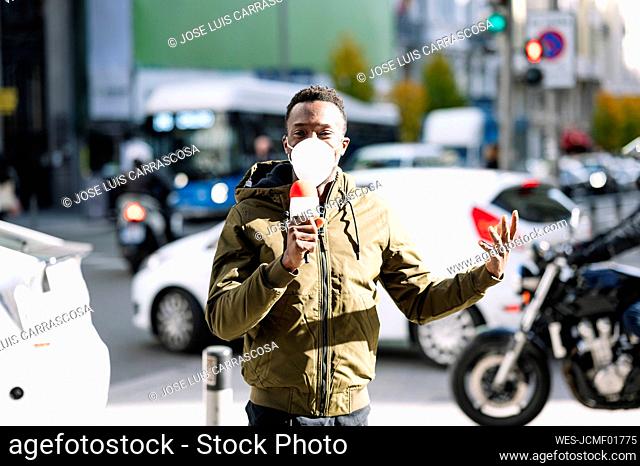 Male TV reporter wearing face mask talking over microphone while standing on street