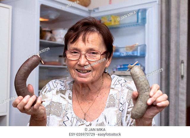 Happy senior woman holding pork liver sausages while standing in front of the open fridge in the kitchen