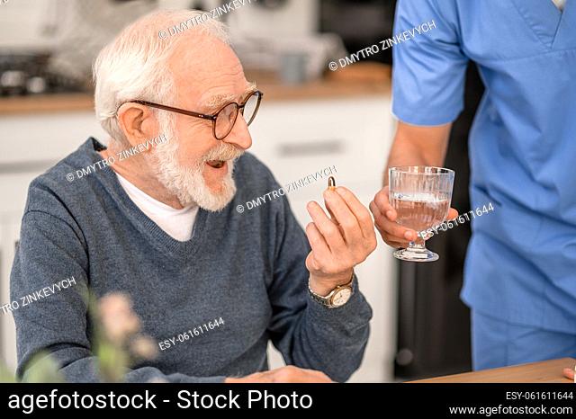 Smiling pensioner examining a pill in his hand while his in-home nurse giving him a glass of water