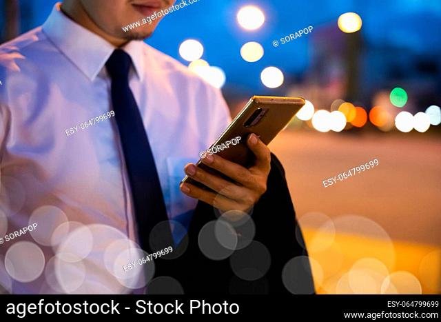 Portrait Asian businessman typing an sms message via smartphone after work near office at night city street, bokeh lights